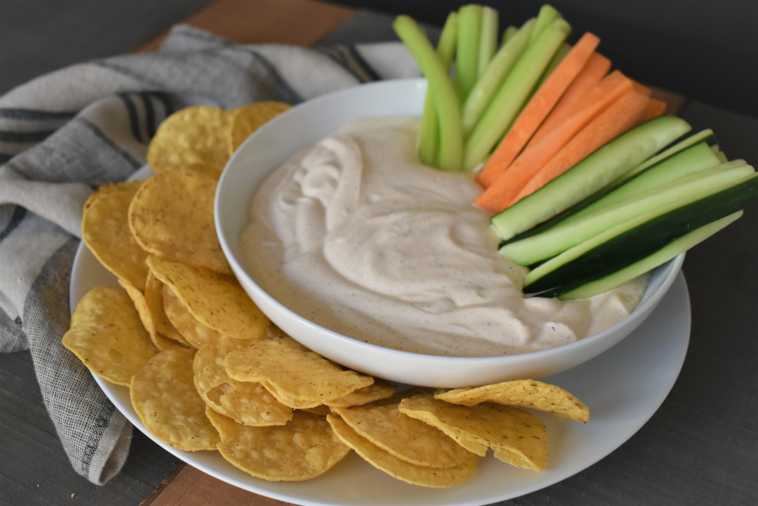 a plate with chips sourrounding a dipping bowl of bbq magic sourn cream dip with celery sticks and other dipping veggies inside