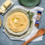 Alt text: A bowl of creamy mashed potatoes topped with butter and pepper, surrounded by butter slices, rosemary, a wooden spoon, and a Meat Magic seasoning bottle.