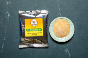 a single silver pouch of chef paul prudhomme's magic glaze next to a pale blue bowl of tan powder on a dark background