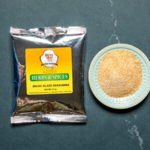 a single silver pouch of chef paul prudhomme's magic glaze next to a pale blue bowl of tan powder on a dark background