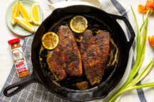 Cast iron skillet with two seasoned fish fillets topped with lemon slices. Nearby are lemon wedges, seasoning, and orange tulips, suggesting a fresh, vibrant meal.