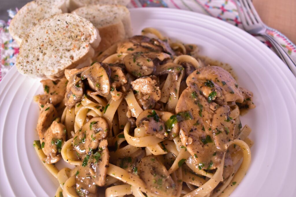 a plate of chicken diane pasta with 2 pieces of garlic bread