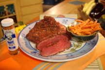 a plate with corned beef brisket with slices cut out, next to a small bowl of seasoned fries on the plate with a container of seasoning magic on the side