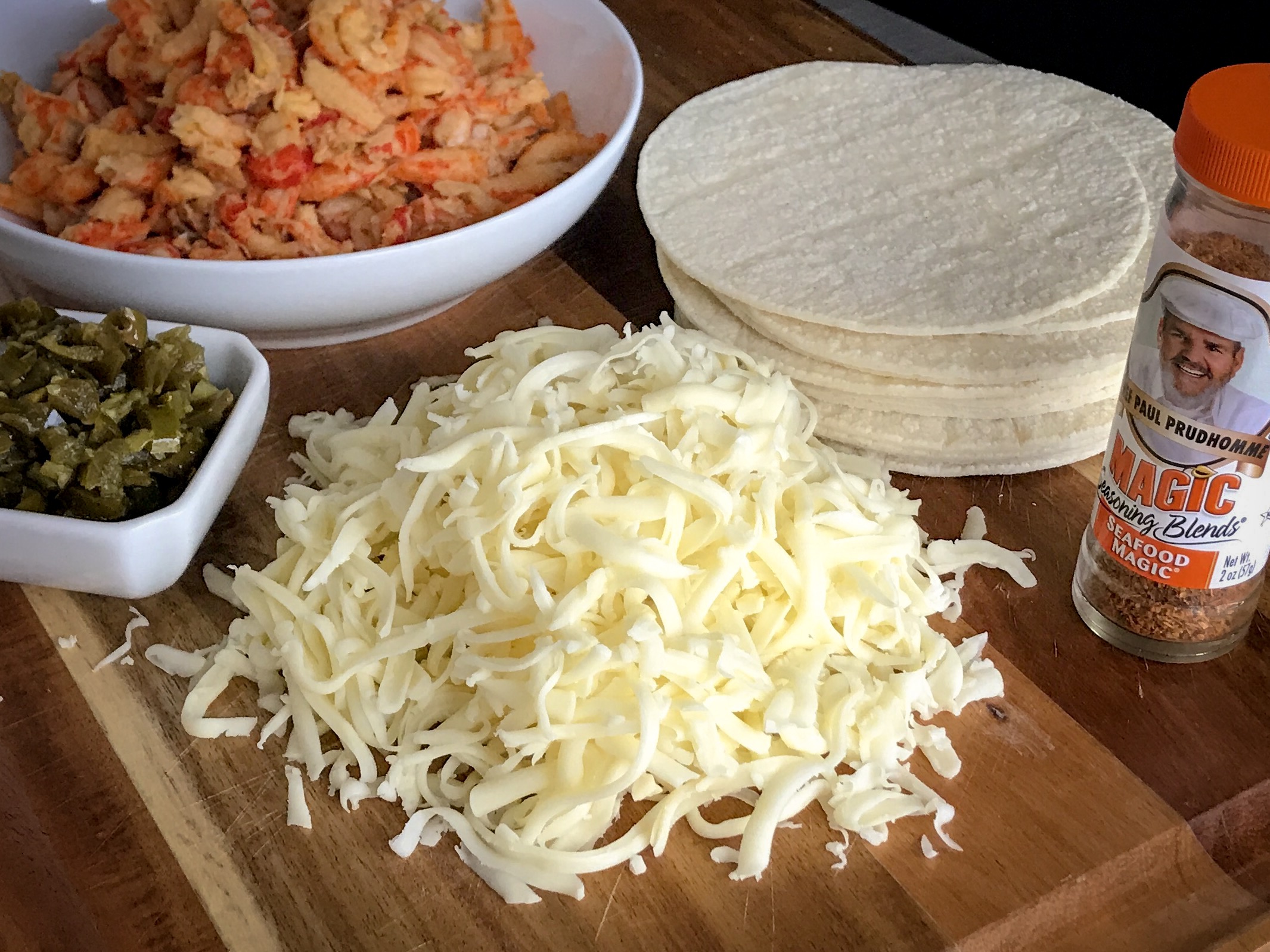 shredded cheese surrounded by a bowl of crawfish, jalapenos, a stack of flour tortillas and a container of seafood magic seasoning blends