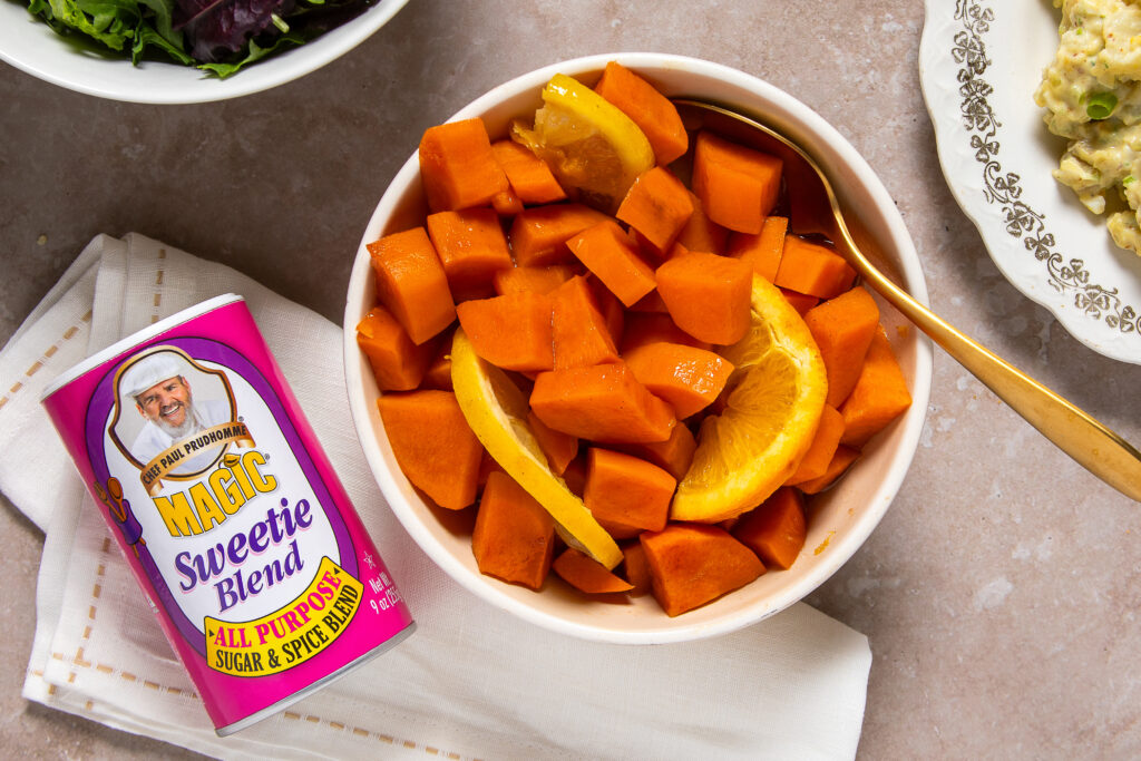 A white bowl of cubed sweet potatoes and orange slices with a gold spoon on a beige surface, next to a can of 'Sweetie Blend' spice resting on a napkin.