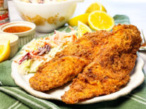 Crispy fried fish fillets served with creamy coleslaw, lemon wedges, and dipping sauce on a white plate.