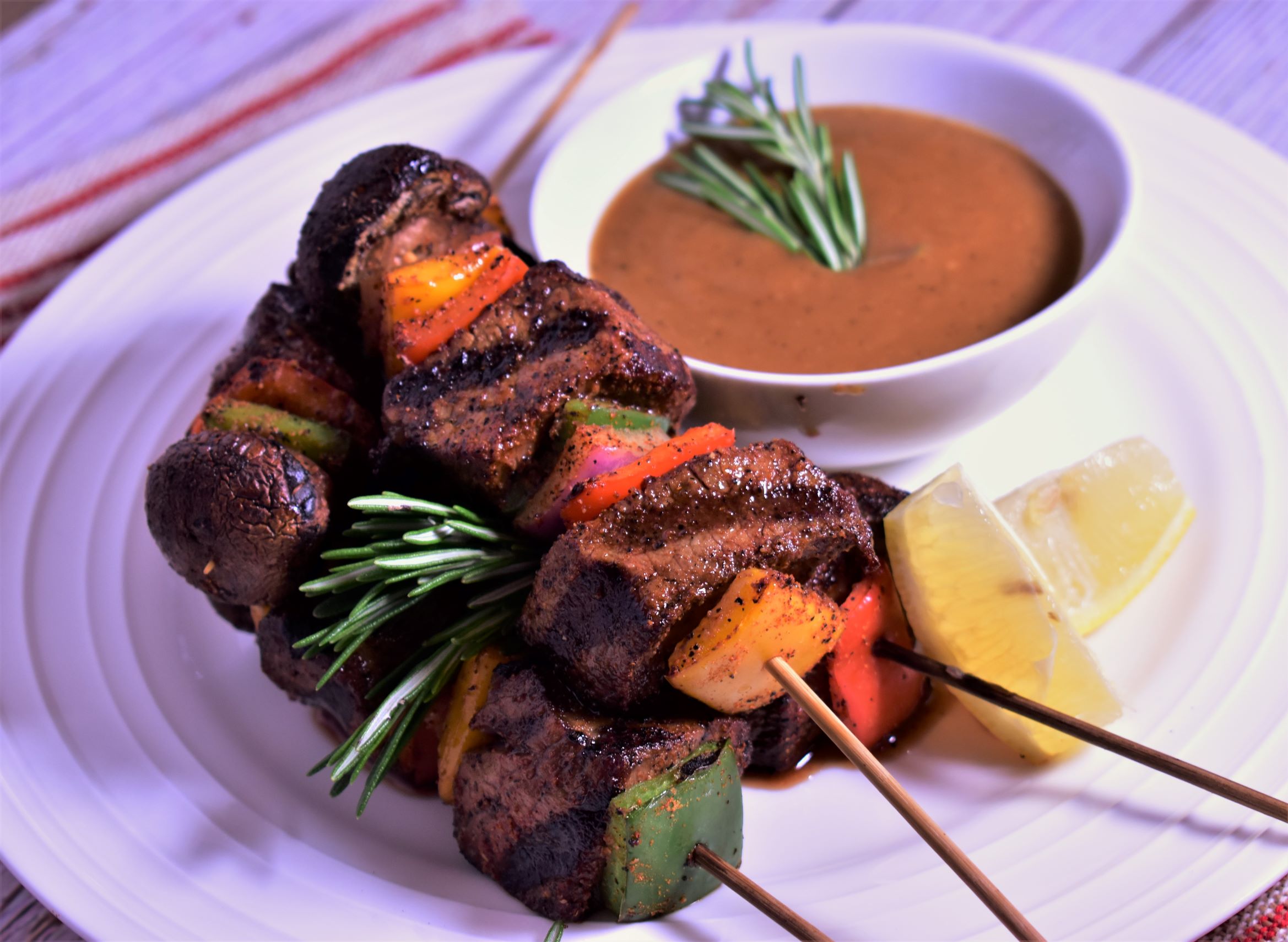 4 rosemary beef skewers on a plate next to 2 lemon slices and a cup of dipping sauce