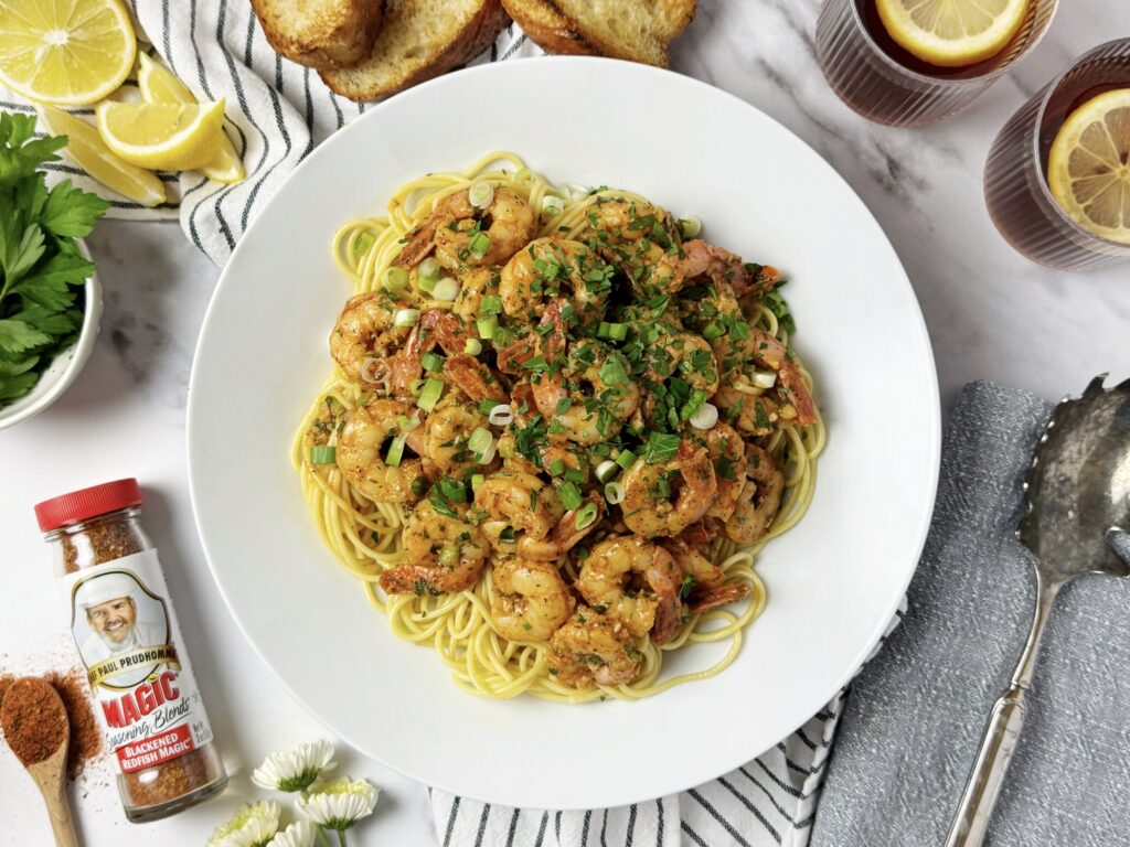 Shrimp scampi with spaghetti on a white plate, garnished with green onions and parsley, served with toasted bread, lemon wedges, and Chef Paul Prudhomme's Magic Blackened Redfish seasoning.