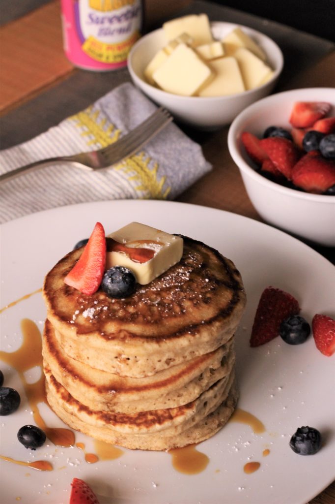 a plate with a stack of 4 fluffy sweetie pancakes topped with a square of butter, a blueberry and a strawberry half