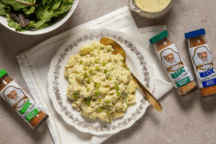 A plate of potato salad drizzled with dressing sits beside a bowl of vibrant greens, surrounded by three bottles of Magic Seasoning.