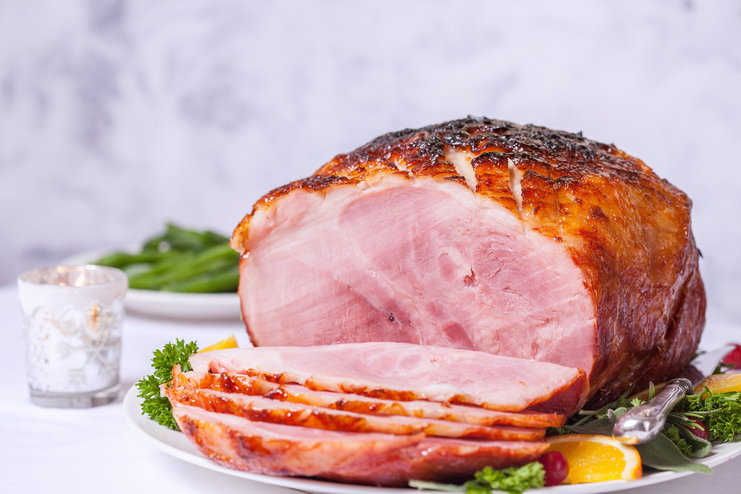 Glazed spiral-cut ham with caramelized exterior, partially sliced on a white platter garnished with fresh parsley, lemon, and green vegetables.