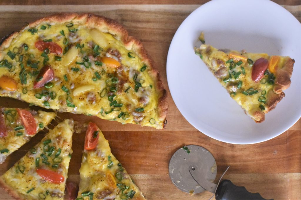 a piece of breakfast pizza on a plate next to the full breakfast pizza the piece was taken from