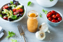 a container of meat magic vinaigrette next to a salad and a bowl of cherry tomates and garlic cloves