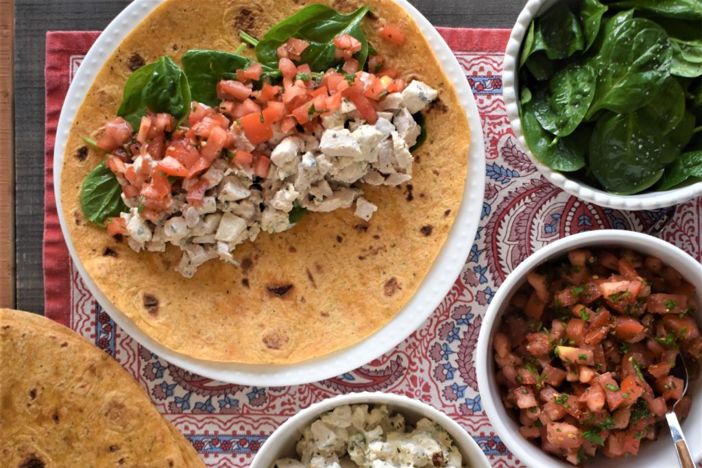 a plate with an italian chicken salad wrap before wrapping up, nnext to bowls with the ingrediants used to make the wrap