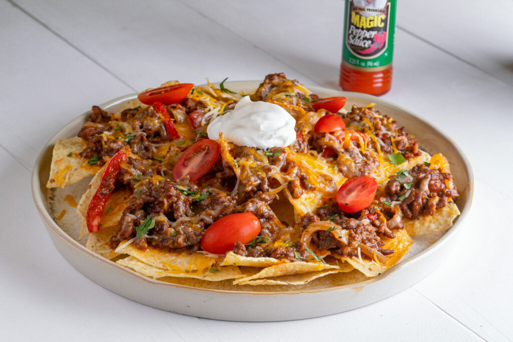 a plate of fajita beef nachos topped with sliced cherry tomatoes and a dollop of sour cream and a bottle of pepper sauce if the background
