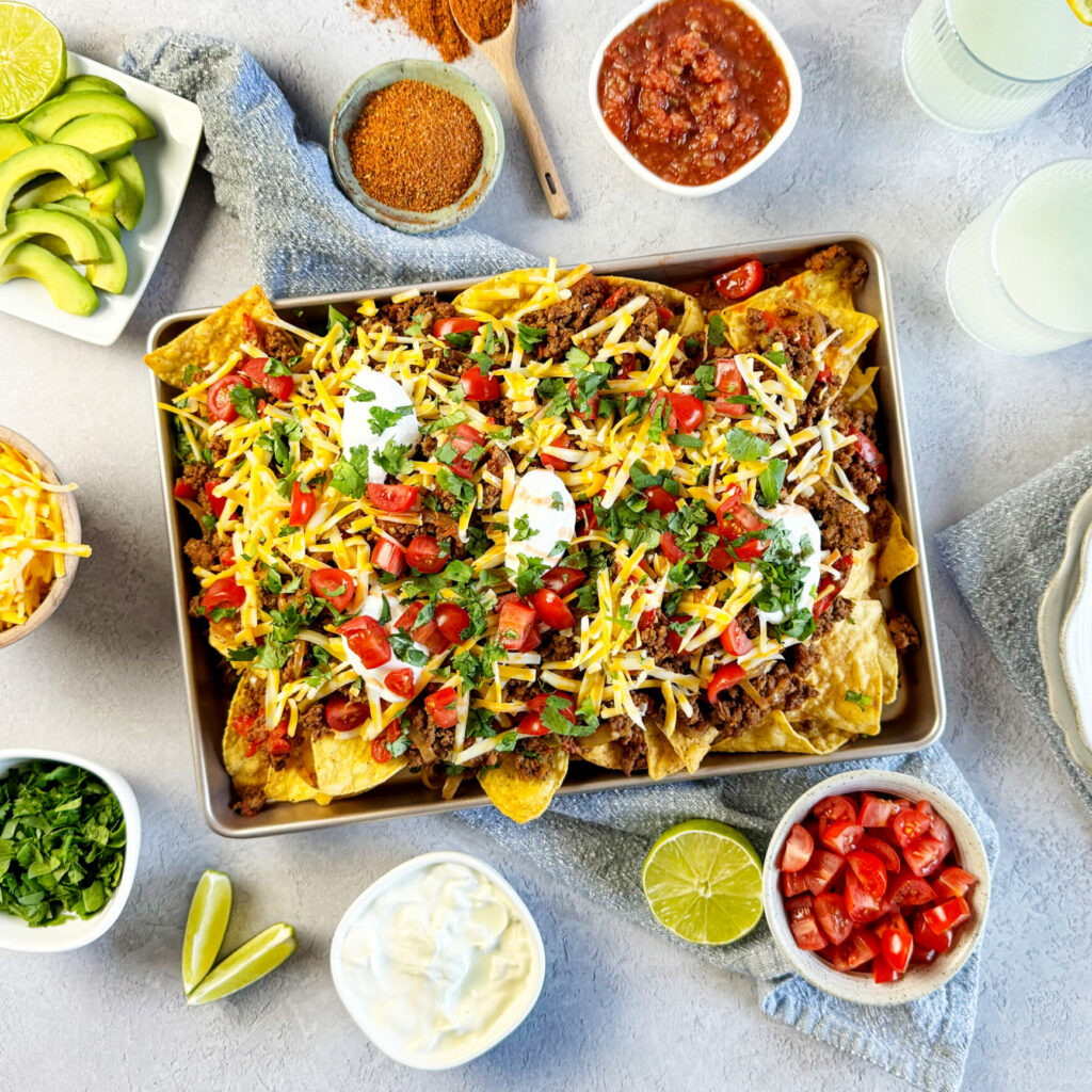 A tray of loaded nachos topped with beef, cheese, tomatoes, sour cream, and cilantro, surrounded by bowls of salsa, shredded cheese, avocado, tomatoes, lime wedges, and drinks with lemon slices.