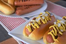 2 chili dogs topped with chopped onions and mustard on a plate next to a another plate with grilled hot dogs and buns