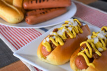Two chili dogs topped with chopped onions and mustard on a plate, next to another plate with grilled hot dogs and buns.