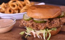 a fried chicken sandwich dressed with lettuce and pickles with a plate of onion rings or curly fries in the background