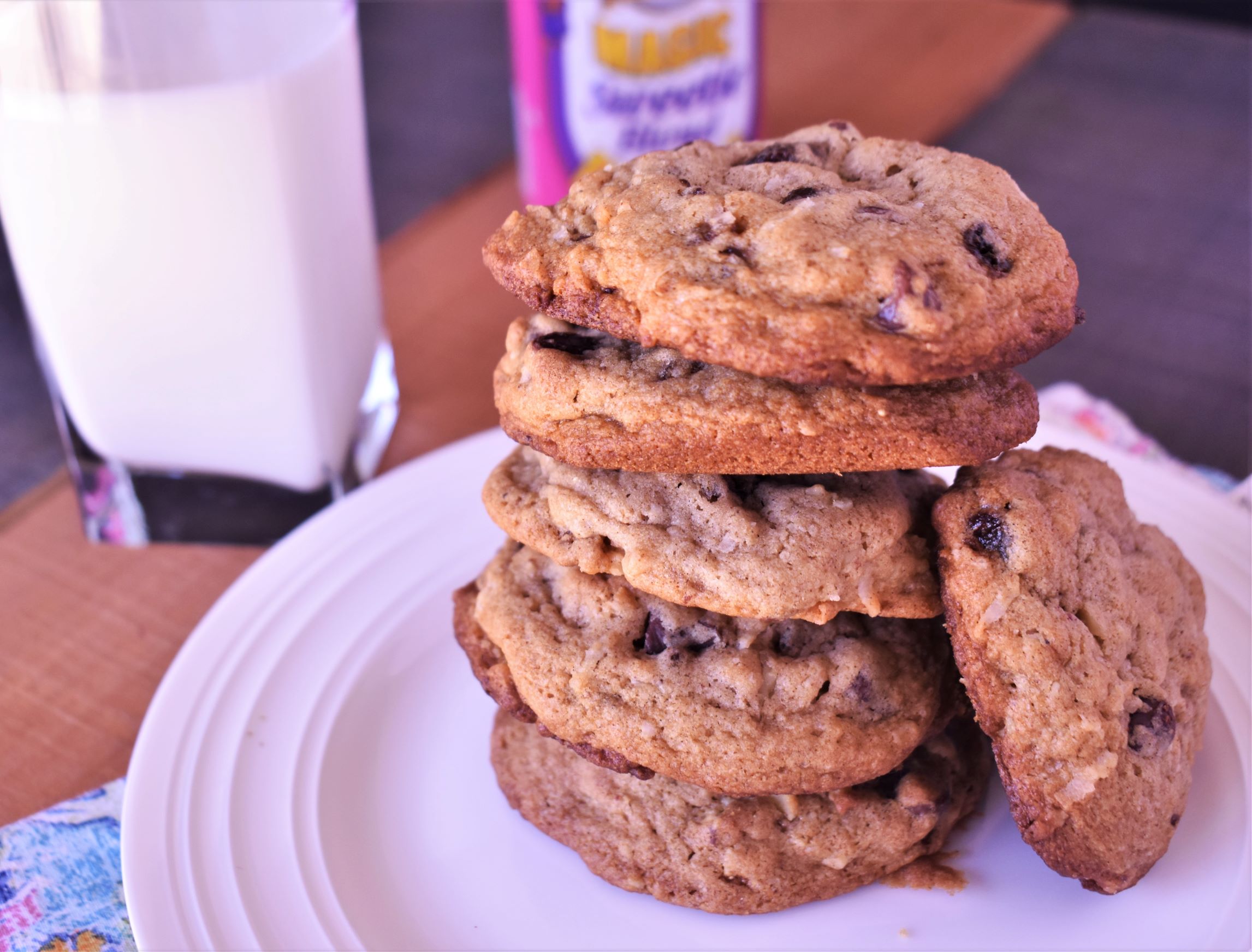 a plate of 6 lori's chocolate chippers stacked on eachother with one on its side, next to a glass of dairy milk