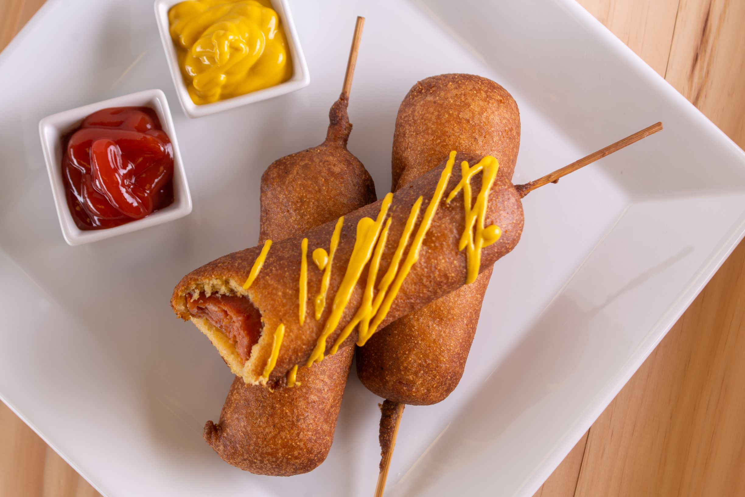 3 andouille corndogs stacked on top of each other with one having a bite taken out of it and drizzled in mustard, next to 2 containers of ketchup and mustard for dipping