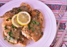 a plate with 3 pieces of chicken piccata topped with a gravy, cilantro and 2 lemon slices