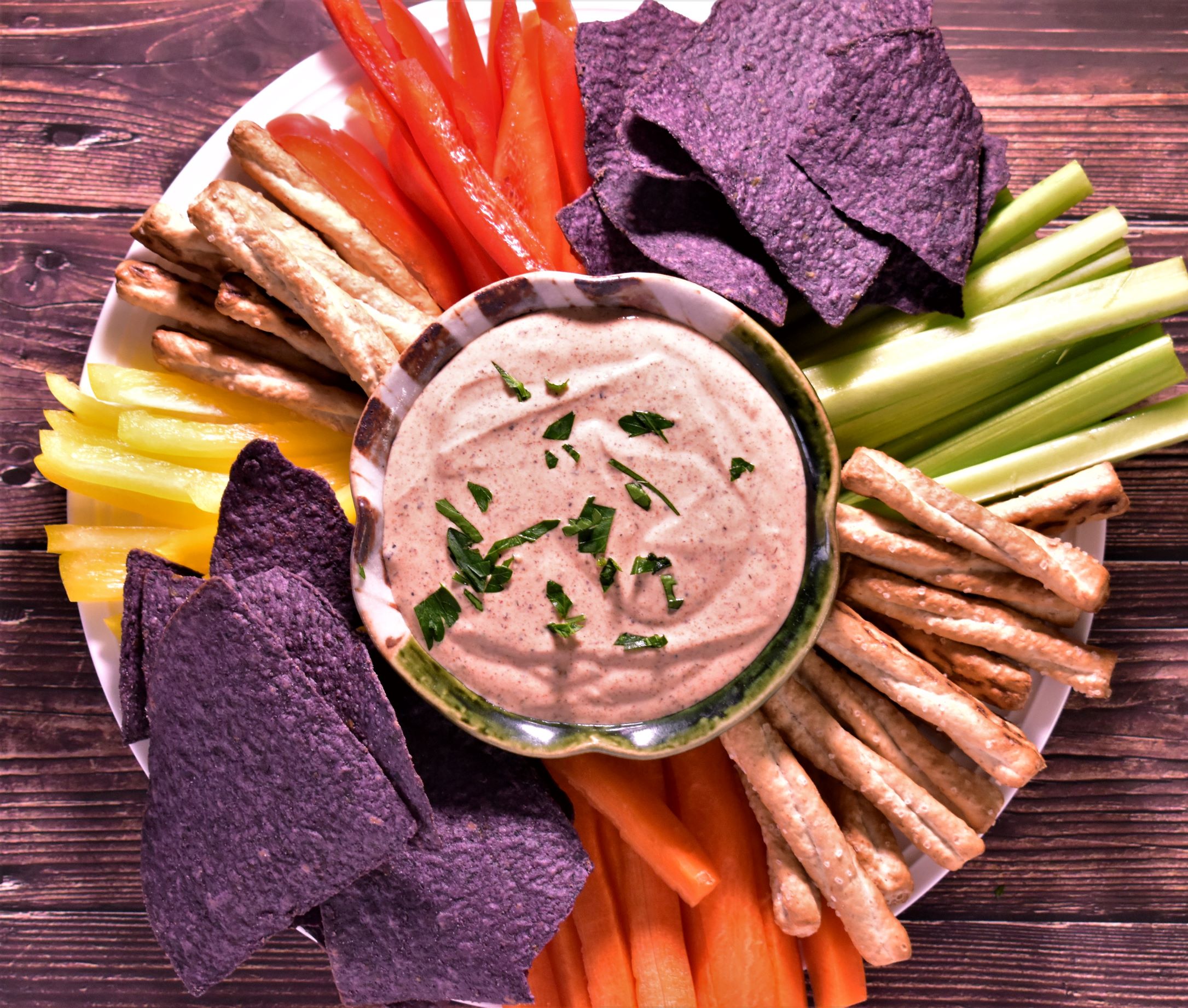 a bowl of greek yogurt chipotle ranch dip surrounded by blue tortilla chips, carrots, celery and other stick shapped dipping snacks