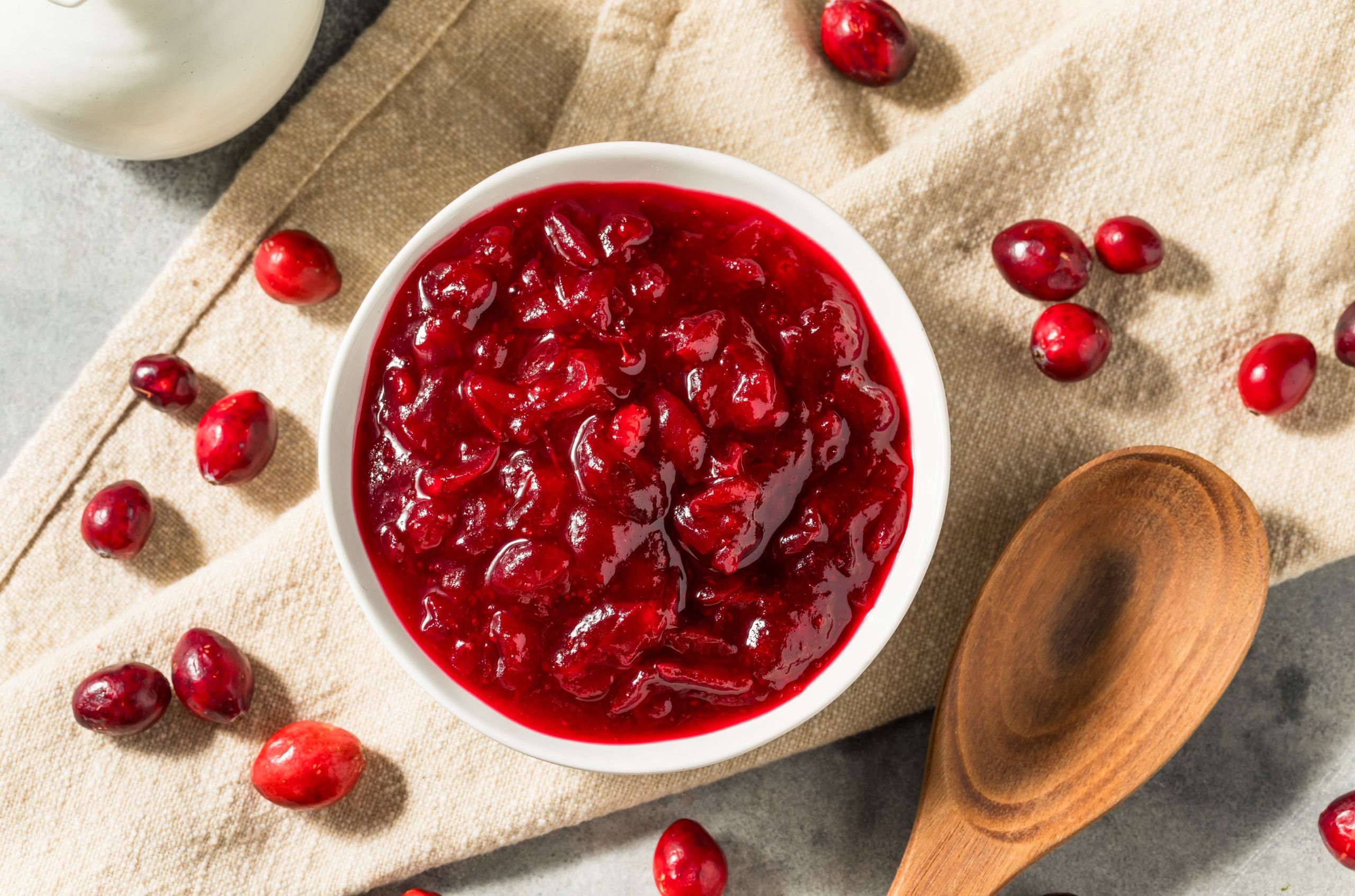 Cranberry Sauce in a white bowl next to a wooden spoon surrounded by cranberries