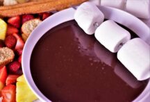 a bowl of chocolate fondue with marshmellows, strawberrys and churros next to it