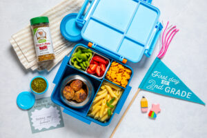 A blue lunchbox with chicken meatballs, penne pasta, strawberries, bell peppers, and crackers, shown with pesto, a seasoning jar, a “First day of 2nd grade” flag, and a card reading “Making new friends!”