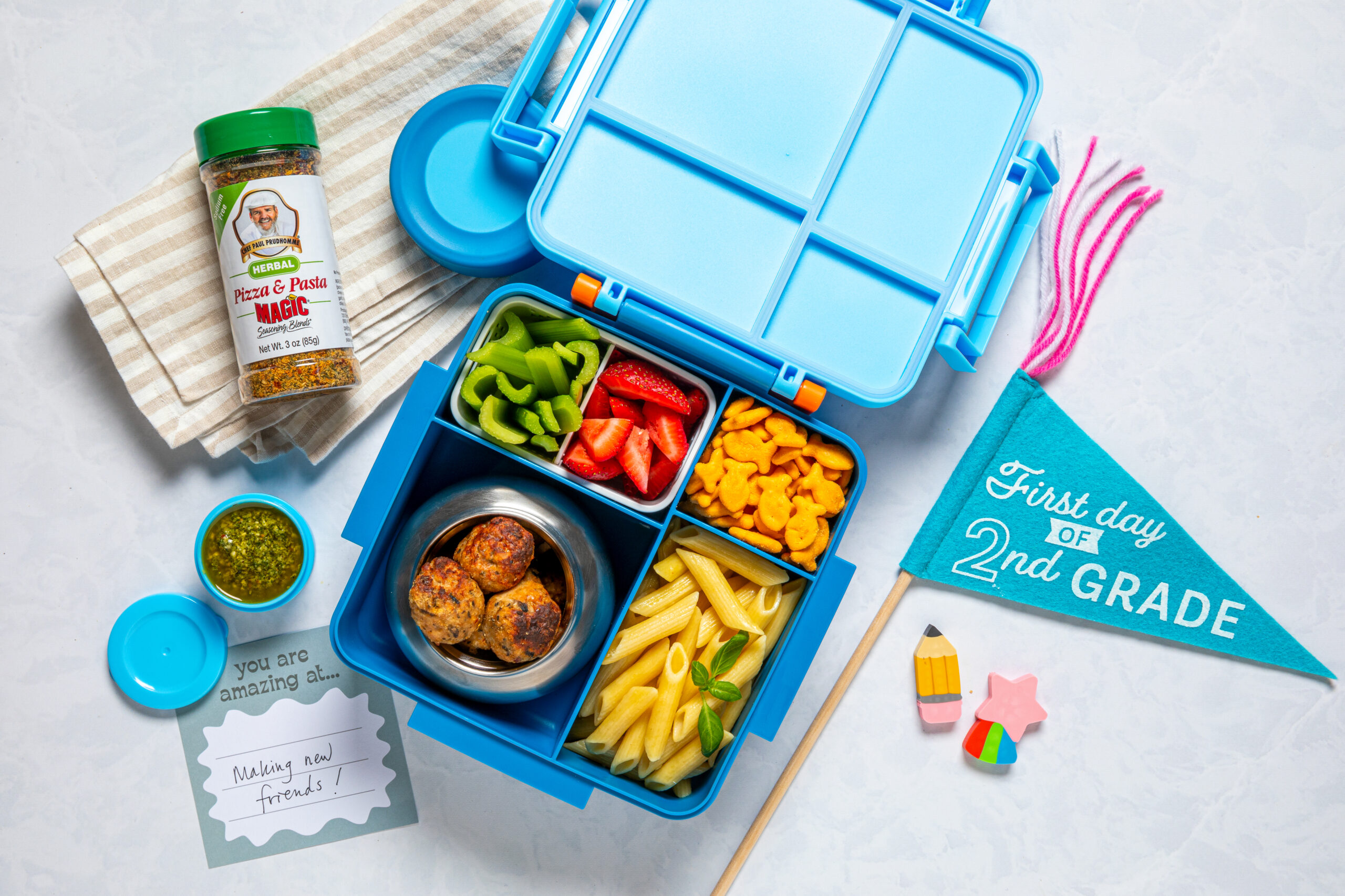 A blue lunchbox with chicken meatballs, penne pasta, strawberries, bell peppers, and crackers, shown with pesto, a seasoning jar, a “First day of 2nd grade” flag, and a card reading “Making new friends!”
