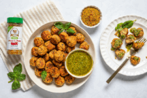 Plate of seasoned meatballs with green pesto dip, garnished with fresh basil. A spice jar and dish of seasoning are nearby on a striped cloth.