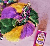 a king cake with a plastic baby and mardi gras beads on it next to a container of magic sweetie blend