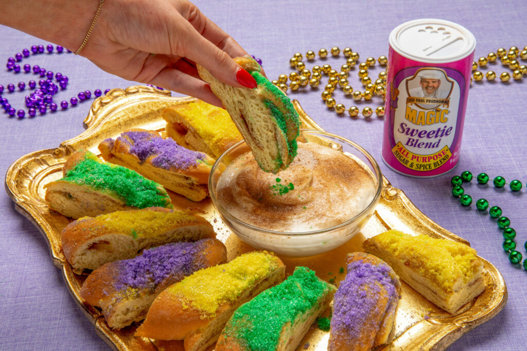someone holding a slice of king cake and dipping it into a bowl of Mardi Gras Sauce next to a gold platter of king cake with a shaker of Sweetie Magic behind it