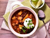 a bowl of chipotle black bean and butternut chilli with a dallop of sour cream and green onions next to a cup of cut limes