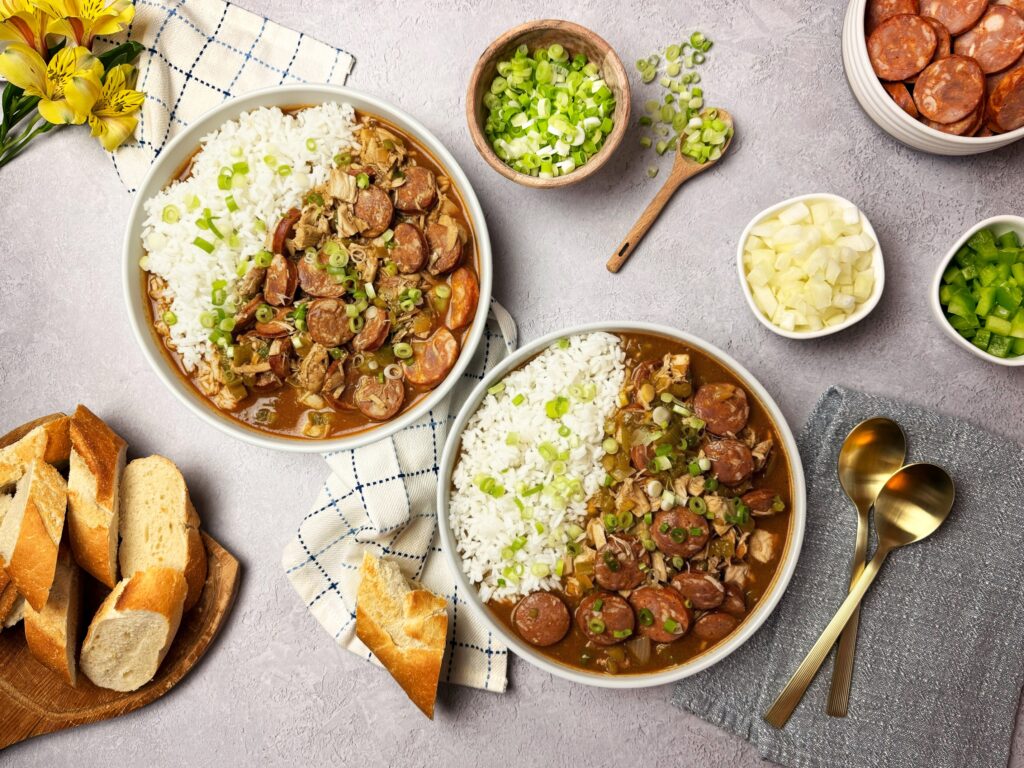 Two bowls of chicken and sausage gumbo with rice, topped with green onions, surrounded by sliced bread, sausage, chopped vegetables, and gold spoons on a gray napkin.