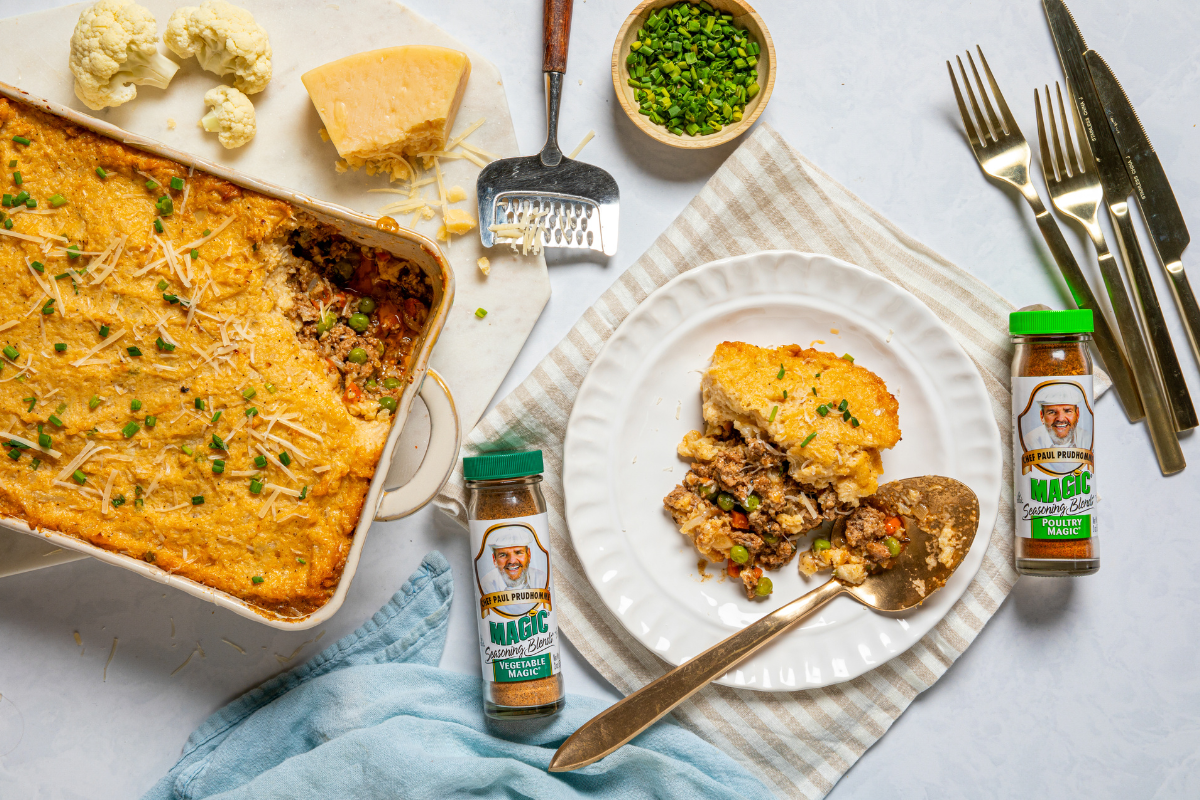 Alt text: A casserole dish of baked shepherd's pie garnished with chives and cheese on a marble surface. Beside it, a plate holds a serving of the pie. Surrounding elements include cheese, cauliflower, a bowl of chopped chives, seasoning bottles, and cutlery, creating a cozy and appetizing scene.