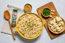 A bowl of mashed cauliflower garnished with chives sits on a table. Surrounding it are raw cauliflower, a spice blend, seasoned crumbs, and a wooden spoon.