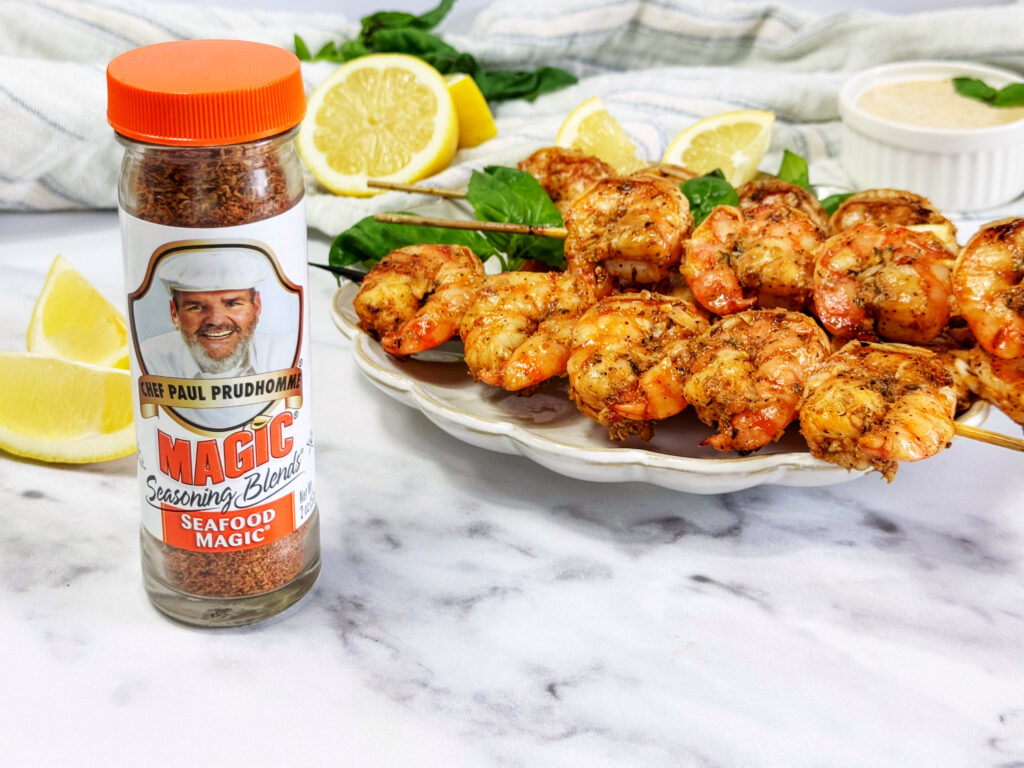 Shrimp Kabobs on a white plate next to a shaker of seafood magic seasoning and lemons