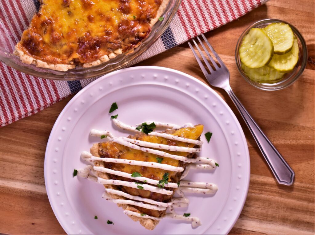 a piece of cheeseburger pie with a white drizzle next to a cup of pickles and the pan of cheeseburger pie the piece came from