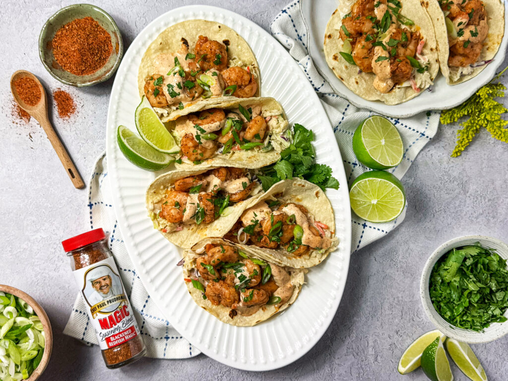 Five Blackened shrimp tacos on a platter topped with creamy sauce and herbs, served with lime wedges and Magic Blackened Redfish seasoning.
