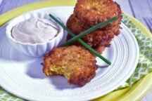 a plate of 4 corn fritters with 3 stacked on top of eachother and 1 with a bite taken out, with a cup of dipping sauce and 2 green onion stalks as garnish