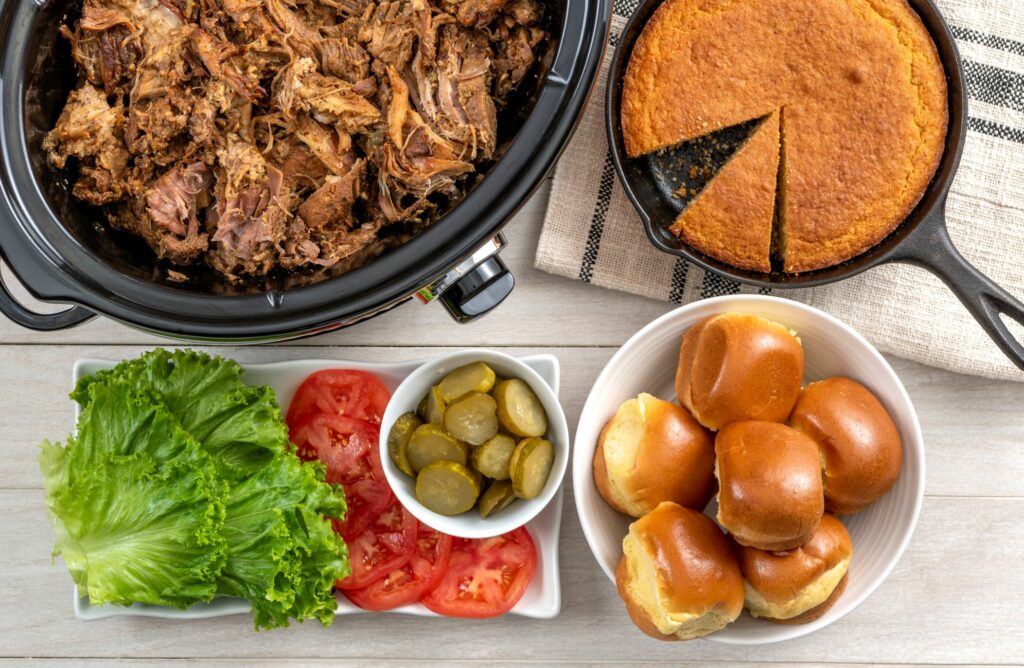 a slow cooker full of slow cooker cooked cochon de lait ,ext to a cast iron skillet of corn bread with a piece taken out, a bowl of rolls and a tray of various fixings inclusing lettuce, pickles and tomatoes