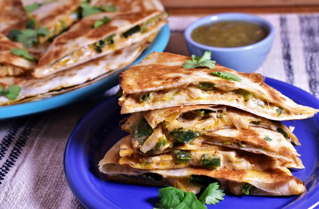 a plate of creamy poblano quesadillas next to a fresh cup of salsa for dipping and a plate of more quesadillas