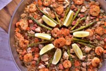 a close up of a big bowl of shrimp, chicken and sausage paella topped with asparagus and lemon slices arranged in a circle