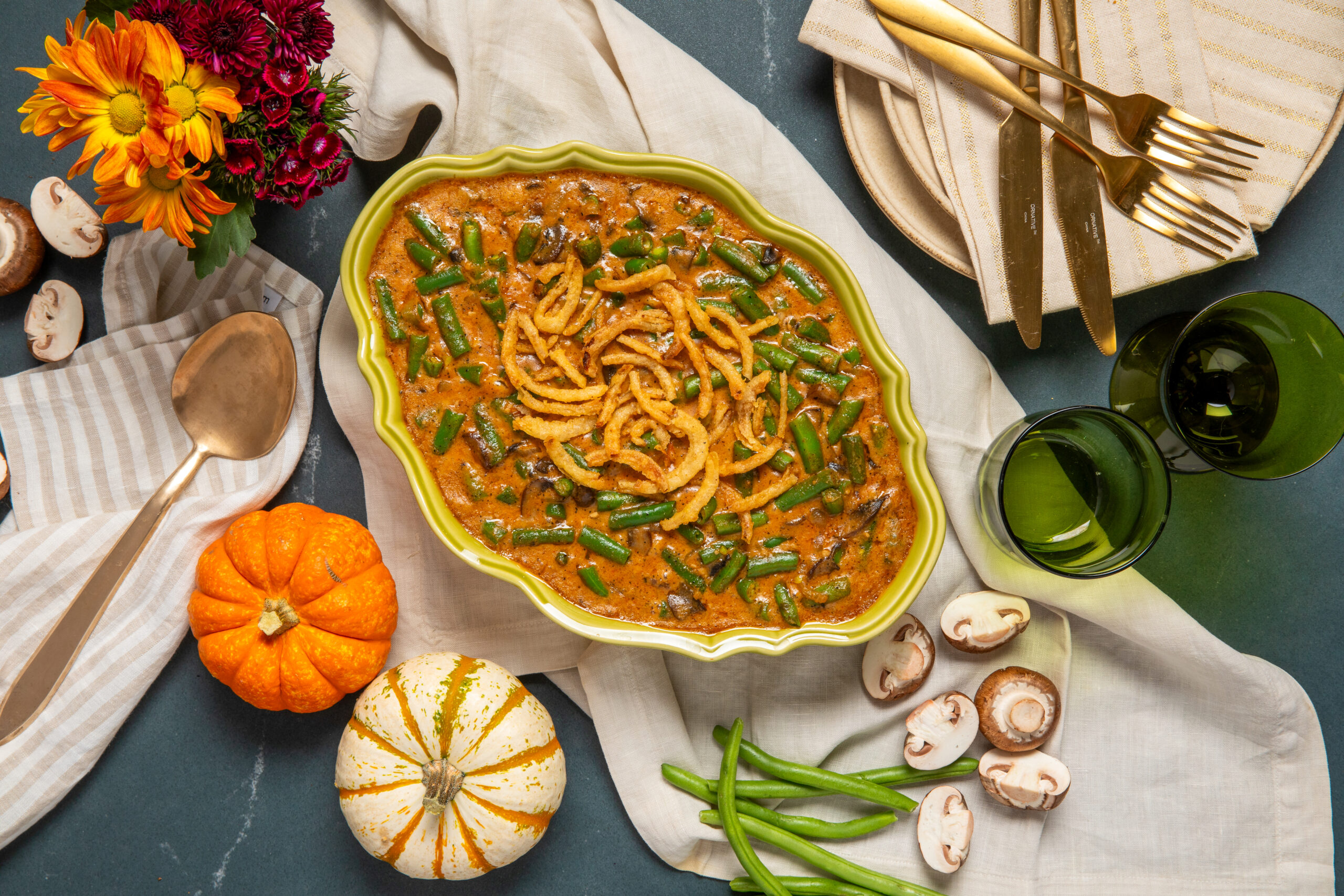 Green Bean Casserole in a white serving dish next to seasonings and pumpkins and plates and forks