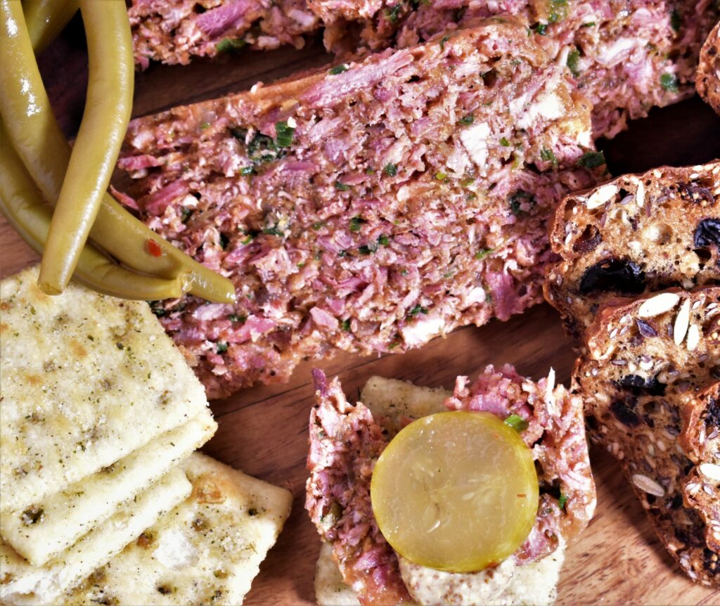 a slice of hoghead cheese next to crackers, green beans, whole grain bread and a pickle slice