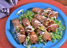 a plate of 5 air fryer chicken taquitos placed on top of a bed a lettuce, drizzled with sour cream and guacamole