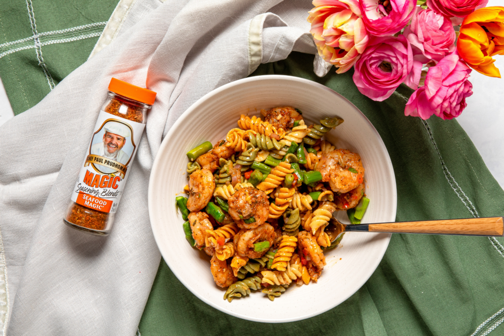 A bowl of colorful rotini pasta with shrimp and green vegetables, garnished with spices. A jar of seasoning and vibrant flowers are beside it on a green cloth.