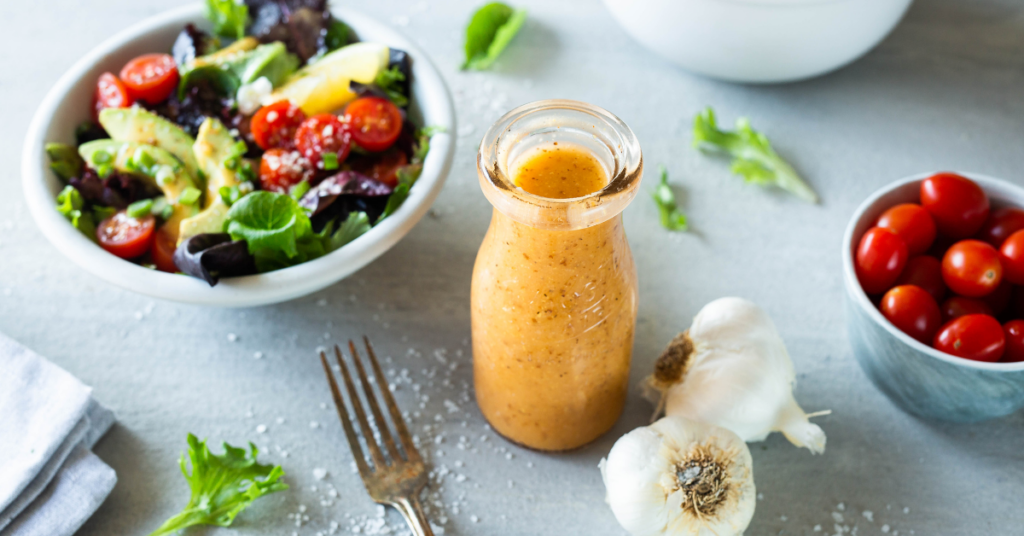 A glass jar of Meat Magic Vinaigrette with a small bowl of salad in the background and a small bowl of cherry tomatoes